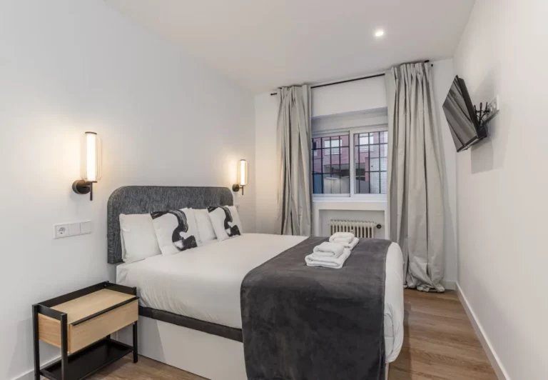 Cama de matrimonio con cabecero tapizado y ventana con cortinas, Queen's Plaza España VI, coliving en Argüelles, Madrid